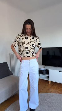 Femme portant un haut à manches courtes fleuri ajustant un pantalon blanc taille haute dans un salon avec une télévision et un canapé.