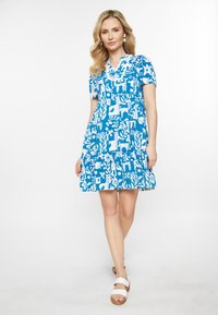Rochie albastră cu modele florale și abstracte albe, mâneci scurte, guler, fustă în straturi și asortată cu sandale albe.
