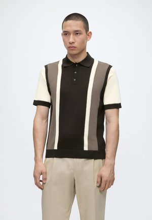 Hombre con camisa polo de manga corta con rayas verticales marrones y crema, combinado con pantalones beige, de pie sobre fondo blanco.