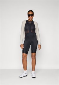 Svart sykkelantrekk med en langermet topp i mesh og tettsittende shorts, kombinert med en kremfarget cardigan og solbriller. Hvite treningssko.