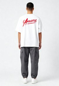 Valge puuvillane T-särk, mille seljal on punane "Amore" tekst, kombineerituna hallide cargo-pükstega ja valgete tossudega.