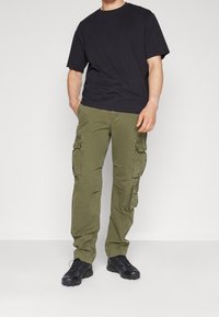 Schwarzes T-Shirt mit kurzen Ärmeln kombiniert mit olivgrünen Cargo-Hosen mit mehreren Taschen und lockerem Schnitt, dazu schwarze Sportschuhe.