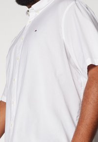 Uomo che indossa una camicia bianca a maniche corte con un piccolo logo ricamato sul petto, mostra il torso superiore e il braccio.
