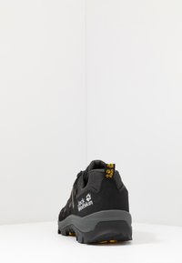 Scarpa da trekking nera con tessuto testurizzato, suola in gomma e accenti gialli. Presenta un tallone rinforzato e un marchio sul retro.