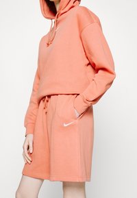 Hooded sweatshirt and shorts set in coral, made of soft fabric; features a front pocket and white Nike logo on both pieces.