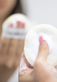Tampon nettoyant blanc rond et texturé avec de la mousse d'un côté, tenu dans une main. L'arrière-plan montre un autre tampon avec des accents rouges.