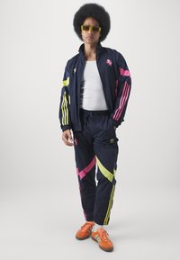 Tracksuit i marineblå med pink og neongule accenter, der har diagonale mønstre. Bæret med orange sko og gule solbriller.
