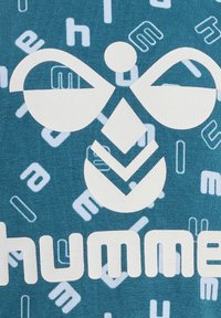 Turkos tyg med en stor vit logotyp som har abstrakta former och bokstäver i ljusare nyanser av blått, vilket skapar en texturerad, lager-på-lager design.