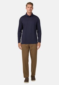 Polo a maniche lunghe blu navy con tre bottoni, abbinato a pantaloni marroni e scarpe marroni, in piedi contro uno sfondo bianco semplice.