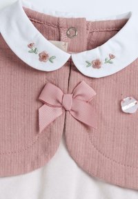 Světlá růžová pletenina s bílým límečkem zdobeným květinovou výšivkou, růžovou mašlí a dekorativním průhledným knoflíkem. Texturovaná látka.