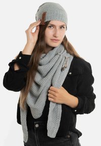 Grijze gebreide beanie met een leren label, gecombineerd met een bijpassende driehoekige sjaal met een gestructureerd patroon en decoratieve hardware accenten.