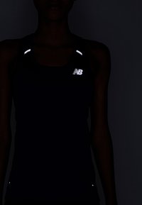 Kvinna som bär en svart sporttanktop med reflekterande New Balance-logotyp och axeldetaljer i svagt ljus.