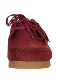 Punainen mokkasiinikenkä, jossa on pyöreä kärki ja nauhat, varustettuna litteällä ruskealla kumipohjalla ja käsinommellulla yksityiskohdalla yläreunassa.