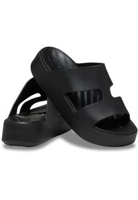 Sandales noires à enfiler avec orteils ouverts, présentant des découpes sur les côtés, un talon épais et une finition mate lisse. Logo visible sur le côté.