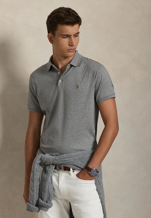 Jeune homme portant un polo gris, un pantalon blanc, une montre bleue et un pull gris noué autour de la taille, regardant sur le côté.