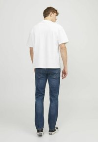 Camiseta blanca de algodón con mangas cortas, combinada con jeans de mezclilla azules. El modelo está de pie con una postura casual, mostrando zapatillas.