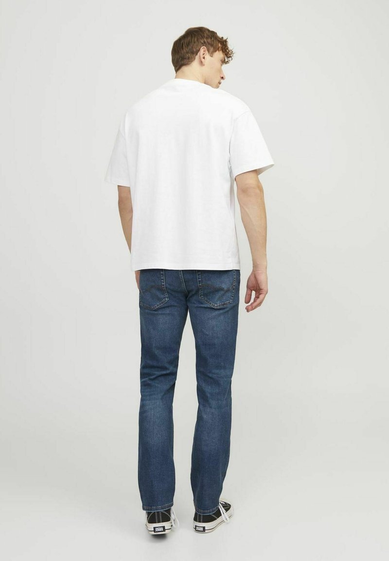 T-shirt en coton blanc à manches courtes, associé à un jean en denim bleu. Le modèle se tient dans une posture décontractée, montrant des baskets.