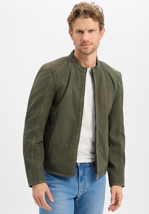 Man met krullend haar die een groene ritsjas draagt over een wit shirt en blauwe spijkerbroek, staand tegen een effen achtergrond.