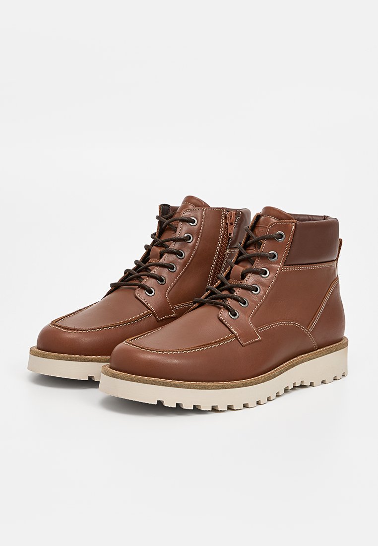 Bottines en cuir marron avec un design robuste, dotées d'un laçage à l'avant, d'une fermeture éclair sur le côté et de semelles en caoutchouc de couleur crème.