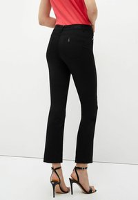 Jeans negros acampanados con una textura suave, cintura de altura media, dos bolsillos traseros y sutiles detalles de costura. Combinados con sandalias negras de tacón alto.