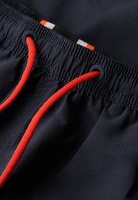 Bañador azul marino con una superficie texturizada, que presenta un cordón de ajuste rojo brillante y detalles en negro. La cinturilla elástica permite ajustar el tamaño.