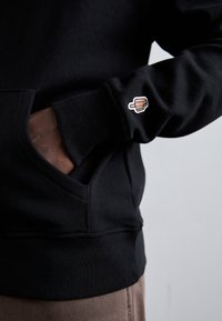 Haut à capuche noir en coton avec une poche frontale, arborant une petite forme de glace brodée sur la manche. Texture douce, coupe décontractée.