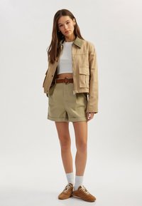 Lichtbeige, kort jasje met een groene kraag, gecombineerd met khaki shorts en bruine sneakers. Model staat tegen een neutrale achtergrond.