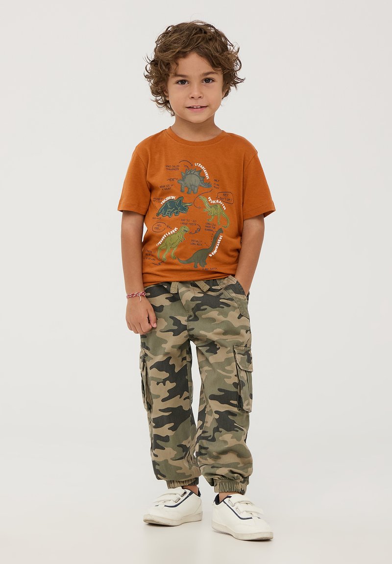 T-shirt arancione in cotone con grafiche di dinosauri e testo, abbinato a pantaloni cargo mimetici verdi con tasche e polsini elasticizzati, indossati con scarpe da ginnastica bianche.