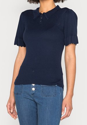 Marineblå strikket bluse med bølget krave, korte pufærmer og knaplukning med tre knapper. Teksturen er ribbet. Båret med denim jeans.