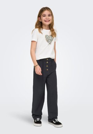 Fille souriante aux longs cheveux portant un t-shirt blanc imprimé de cœurs, un pantalon noir ample avec des boutons, des baskets noires et blanches, et un bracelet.
