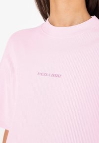 Ružový rebrovaný krátky rukáv tričko s vysokým golierom a vyšitým logom "PEGADOR" v kontrastnej farbe. Mäkká textúra tkaniny.