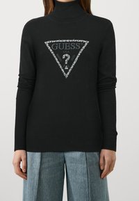 Personne portant un pull col roulé noir avec un logo en triangle scintillant de la marque Guess et un jean large bleu, se tenant devant un fond uni.