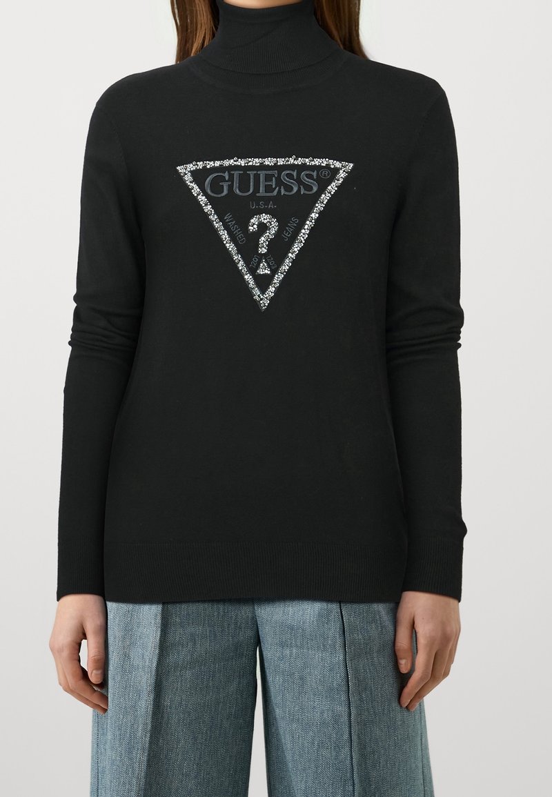 Personne portant un pull col roulé noir avec un logo en triangle scintillant de la marque Guess et un jean large bleu, se tenant devant un fond uni.