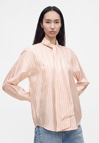 Femme aux longs cheveux noirs portant un chemisier rayé rose clair avec un détail cravate au col et un jean bleu, debout les mains sur les hanches sur fond blanc.