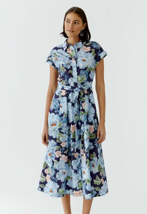 Femme portant une robe midi bleu marine avec un grand imprimé floral bleu clair et rose, manches courtes, boutonnière devant et ceinture nouée à la taille.