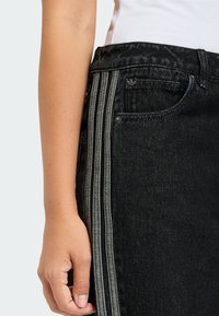 Τα μαύρα τζιν από τζιν διαθέτουν τρεις κάθετες γκρι ρίγες κατά μήκος της πλευράς, με κλασικό σχέδιο πέντε τσεπών, συμπεριλαμβανομένης μιας μικρής μπροστινής τσέπης.