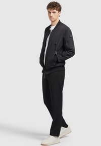 Schwarze Bomberjacke mit Reißverschlüssen, gerippten Bündchen und einem Patch am Ärmel. Über einem weißen Shirt getragen, kombiniert mit schwarzen Hosen und weißen Sneakers.