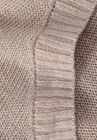 Pletený svetr v světle béžové barvě. Na levé straně má texturovaný vzor a na pravé straně žebrovaný okraj, zobrazující různé techniky pletení.
