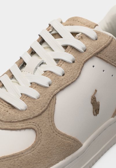 Pantof sport bej și alb cu panouri din piele întoarsă texturată, șireturi albe și un mic logo în relief cu un cal și un călăreț pe lateral.