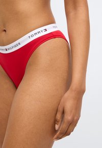 Rød bikini med et hvidt elastikbånd med "TOMMY HILFIGER" logo. Glat tekstur med lavtaljet snit og minimal dækning.