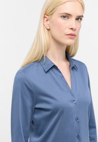 Lys blå langærmet skjorte med krave, der har en knaplukning fortil og blødt, glat stof. Ingen synlige mønstre eller detaljer.