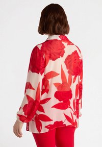 Blouse en dentelle à manches longues crème avec un imprimé floral rouge surdimensionné, présente une coupe décontractée, un col et des poignets élastiqués.