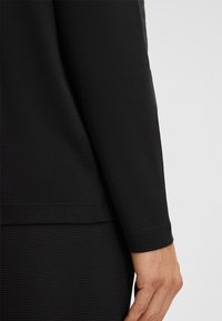 Czarna koszula z długim rękawem o gładkiej, matowej tkaninie. Charakteryzuje się luźnym krojem, mankietami bez zapięć oraz prostym dołem.