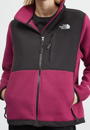 Kvinde iført en lilla og sort The North Face fleecejakke med lynlåslommer og logo på brystet, hænderne i sidelommerne.