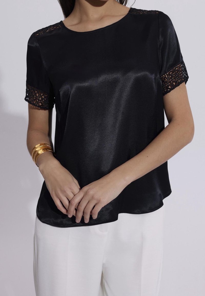 Femme portant un chemisier noir en satin à manches courtes avec bordure en dentelle et un pantalon blanc, accessoirisée de trois bracelets en or au poignet gauche.