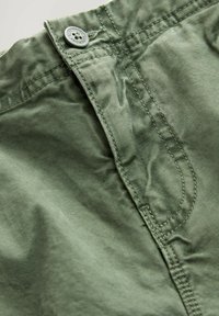 Pantalones cortos verdes de algodón con cierre de botón, costuras visibles y una textura ligeramente arrugada. Primer plano del detalle de la cintura.