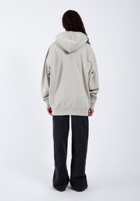 Sudadera oversized gris claro con una gran capucha y puños acanalados, combinada con pantalones de pierna ancha oscuros. Tela suave, corte relajado, diseño minimalista.