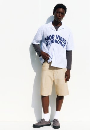Junger Mann trägt ein weißes "Good Vibes Laundromat" Hemd über einem schwarzen Langarmtop, beige Shorts, weiße Socken und graue Turnschuhe und steht vor einer weißen Wand.