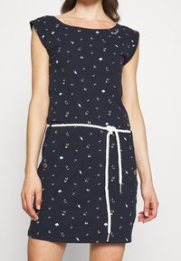 Granatowa sukienka z krótkimi rękawami, występująca w wzór składający się z jasnoniebieskich i białych kształtów. Zawiera białą sznurówkę w talii oraz drewniane akcenty w postaci guzików.