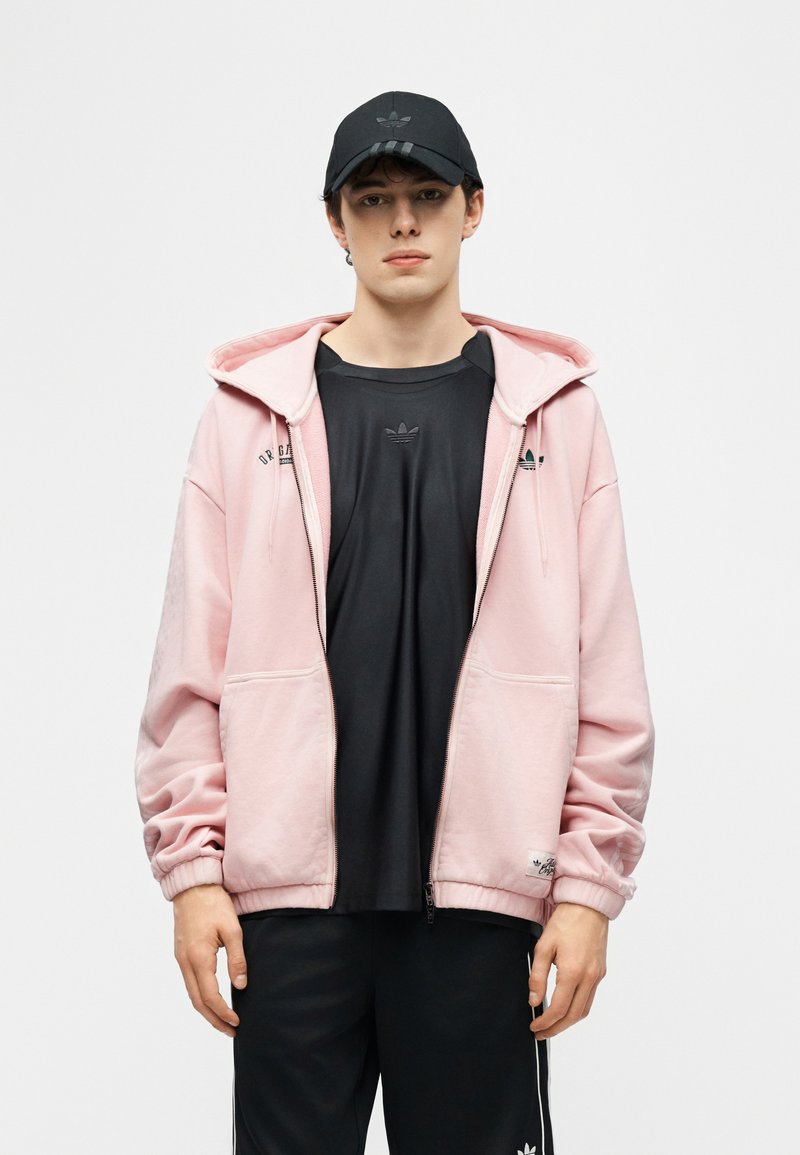 Jeune homme portant une casquette noire, un t-shirt Adidas noir, un sweat à capuche zippé rose clair et un pantalon Adidas noir sur un fond uni.